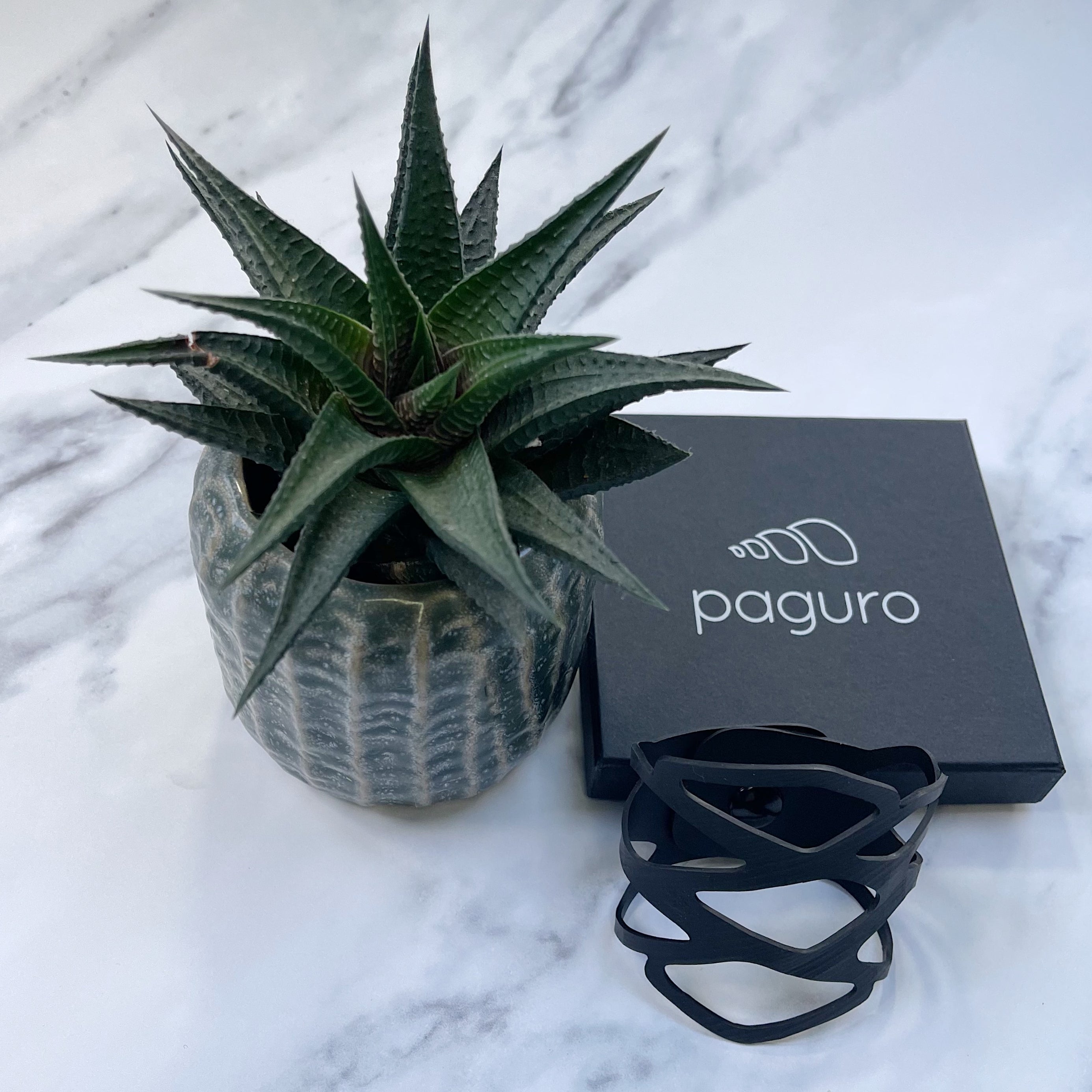 Black recycled rubber bracelet by Paguro Upcycle displayed next to plant and branded box