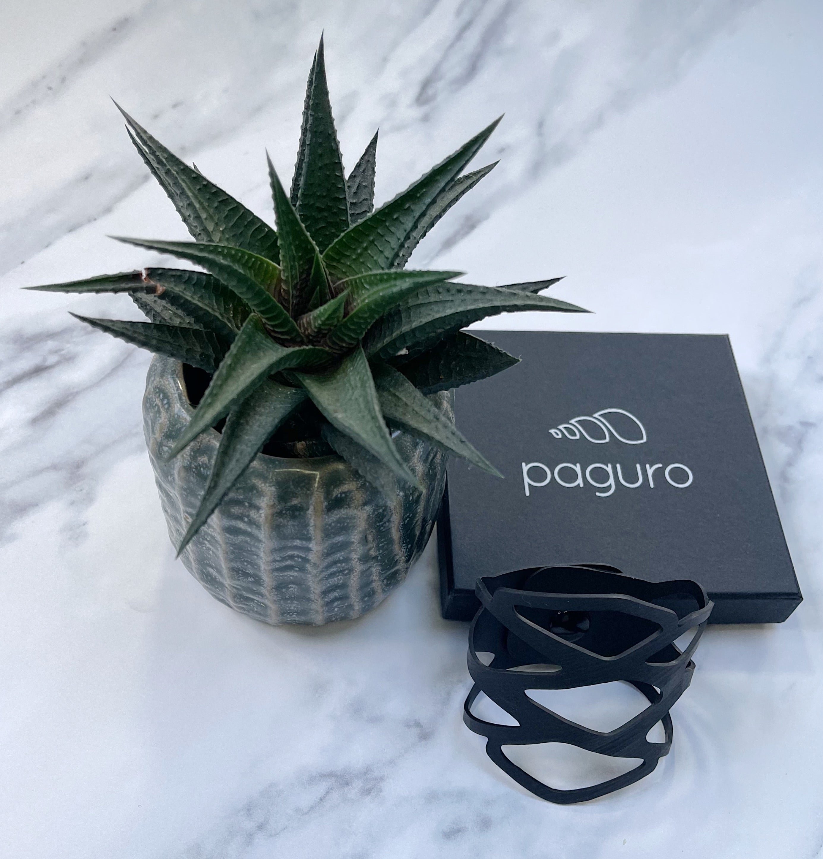 Black recycled rubber bracelet by Paguro Upcycle displayed next to plant and branded box